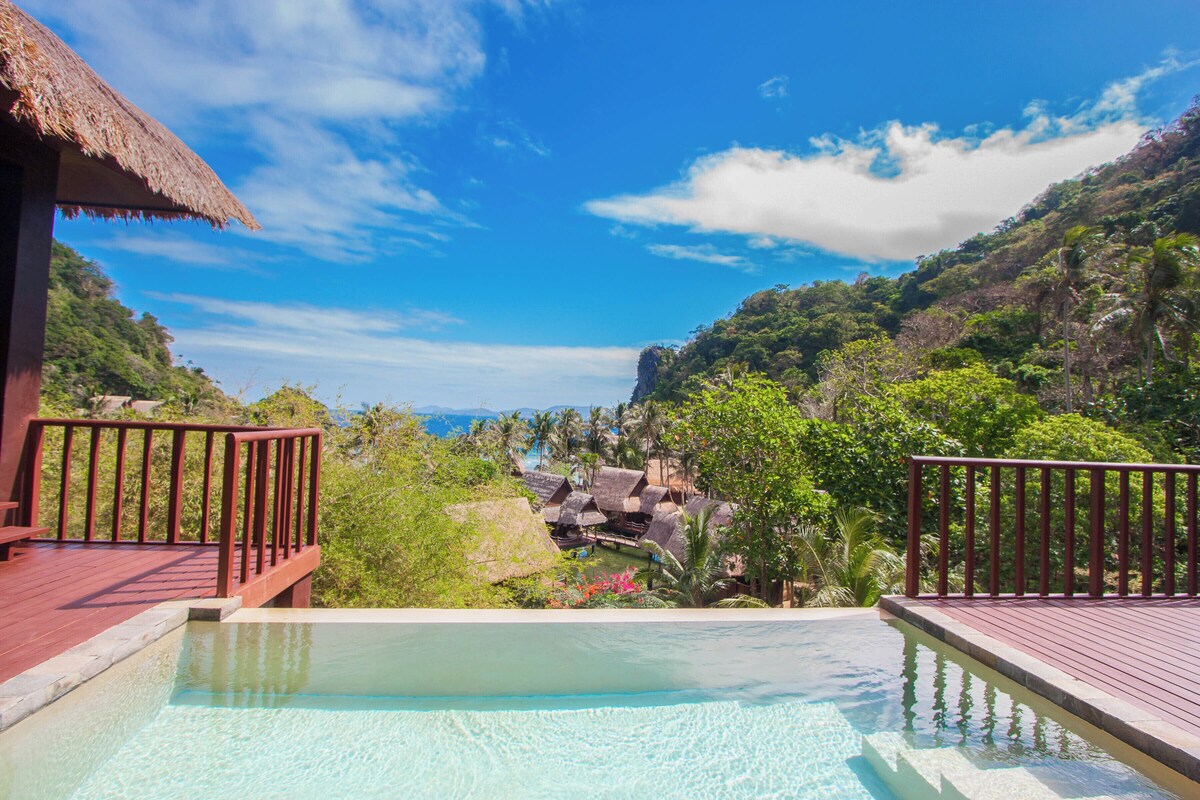 A pool with a wooden deck and a view of a tropical resort with thatched-roof buildings and lush greenery.