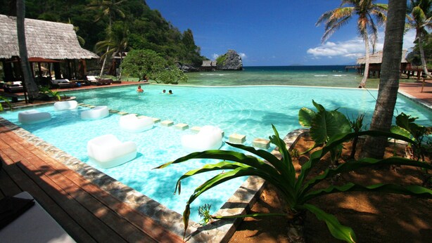 A clear blue pool with floating seats, surrounded by palm trees and a mountain backdrop. - Cauayan Boutique Private Island (El Nido)
