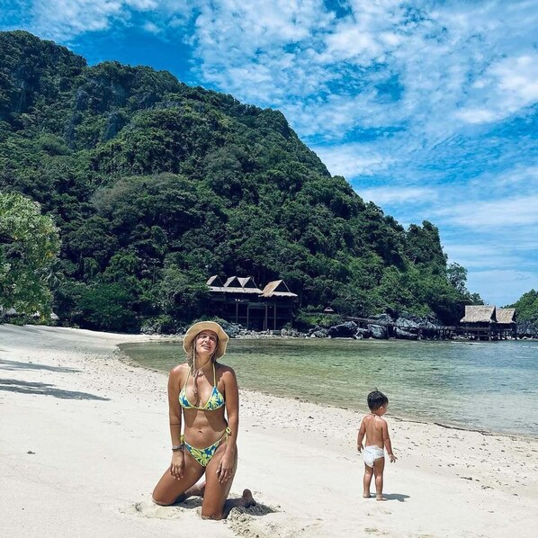A beach with a woman in a bikini and a child, a thatched-roof structure, and lush green hills in the background.