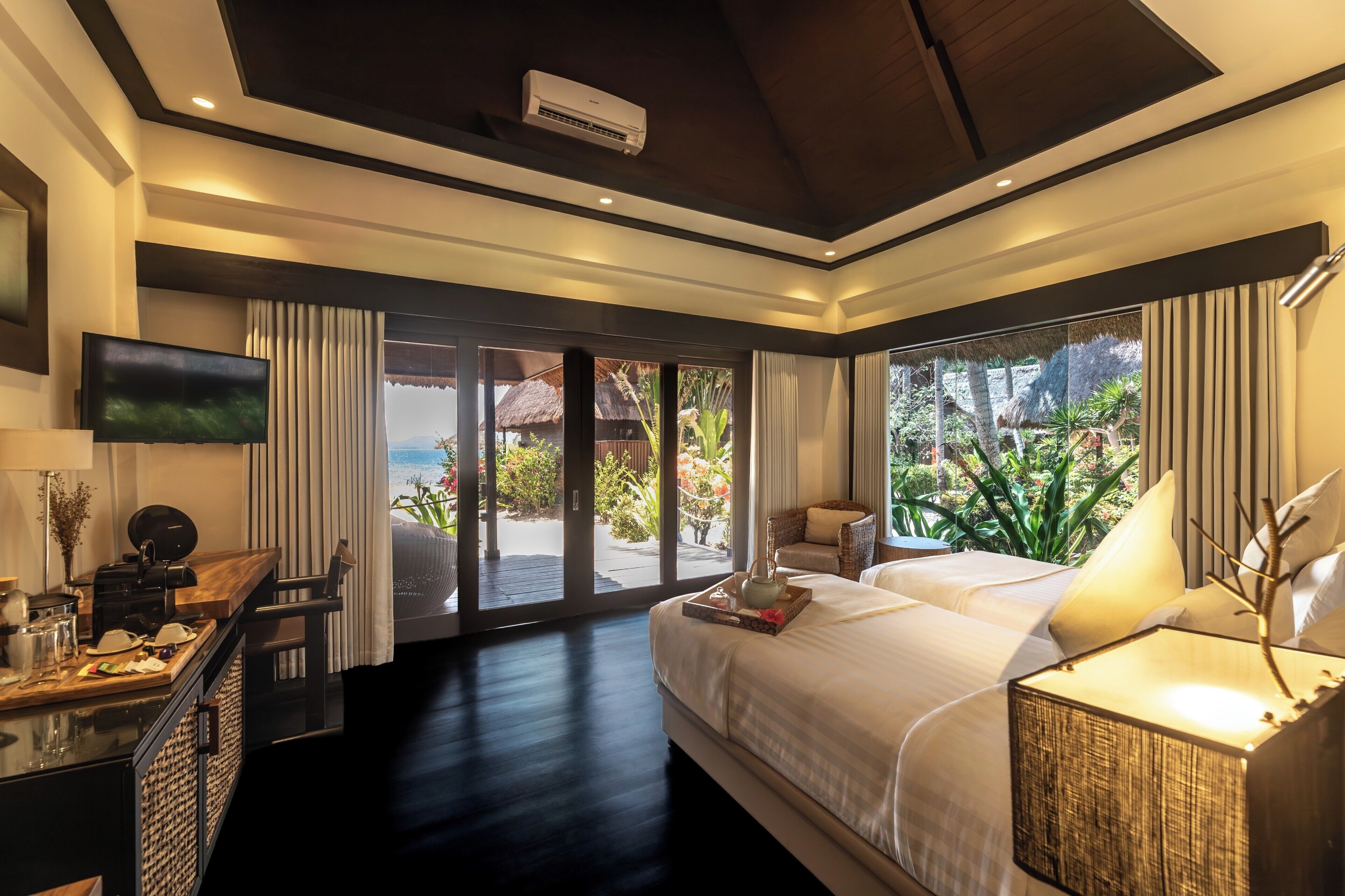 A hotel room with a bed, a desk with a coffee maker, a TV, and a view of a garden and beach through large windows.