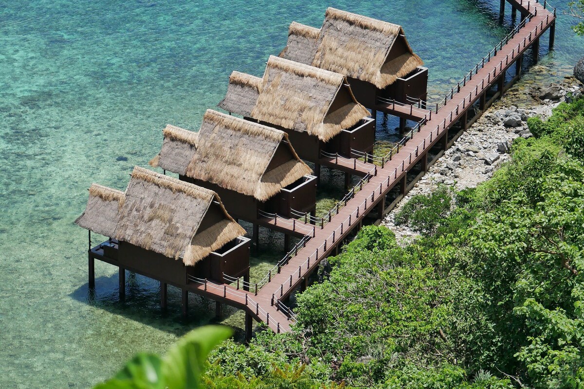 Overwater bungalows with thatched roofs connected by a wooden walkway extending into clear blue water.
