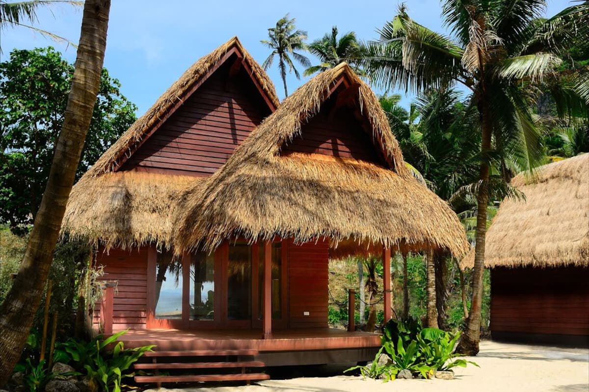 Ein zweistöckiges Reetdachbungalow mit Holzfassade, umgeben von Palmen und einem Sandstrand.