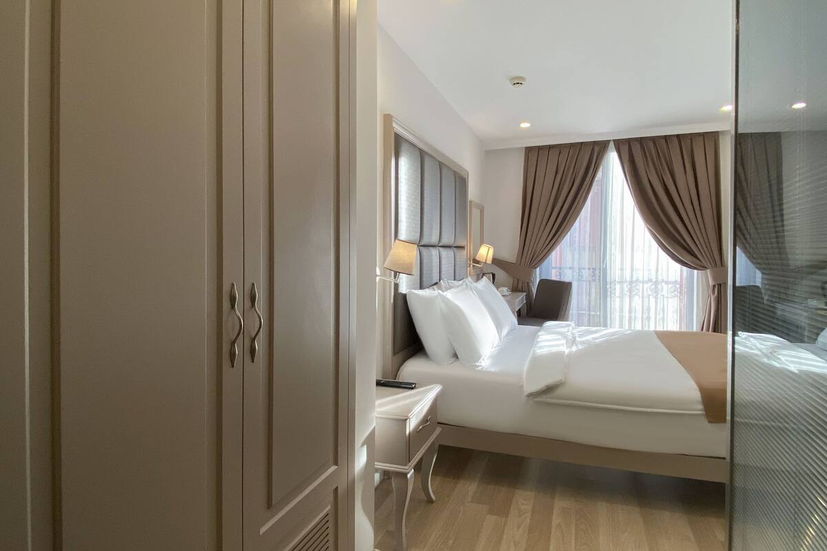 Une chambre d’hôtel moderne avec un grand lit, un bureau et une chaise. Il y a une fenêtre avec des rideaux, un miroir et une lampe fixée au mur.