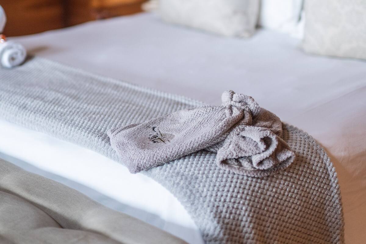 A neatly made bed with a grey blanket and a folded towel, a white pillow, and a tufted headboard in a wooden room.