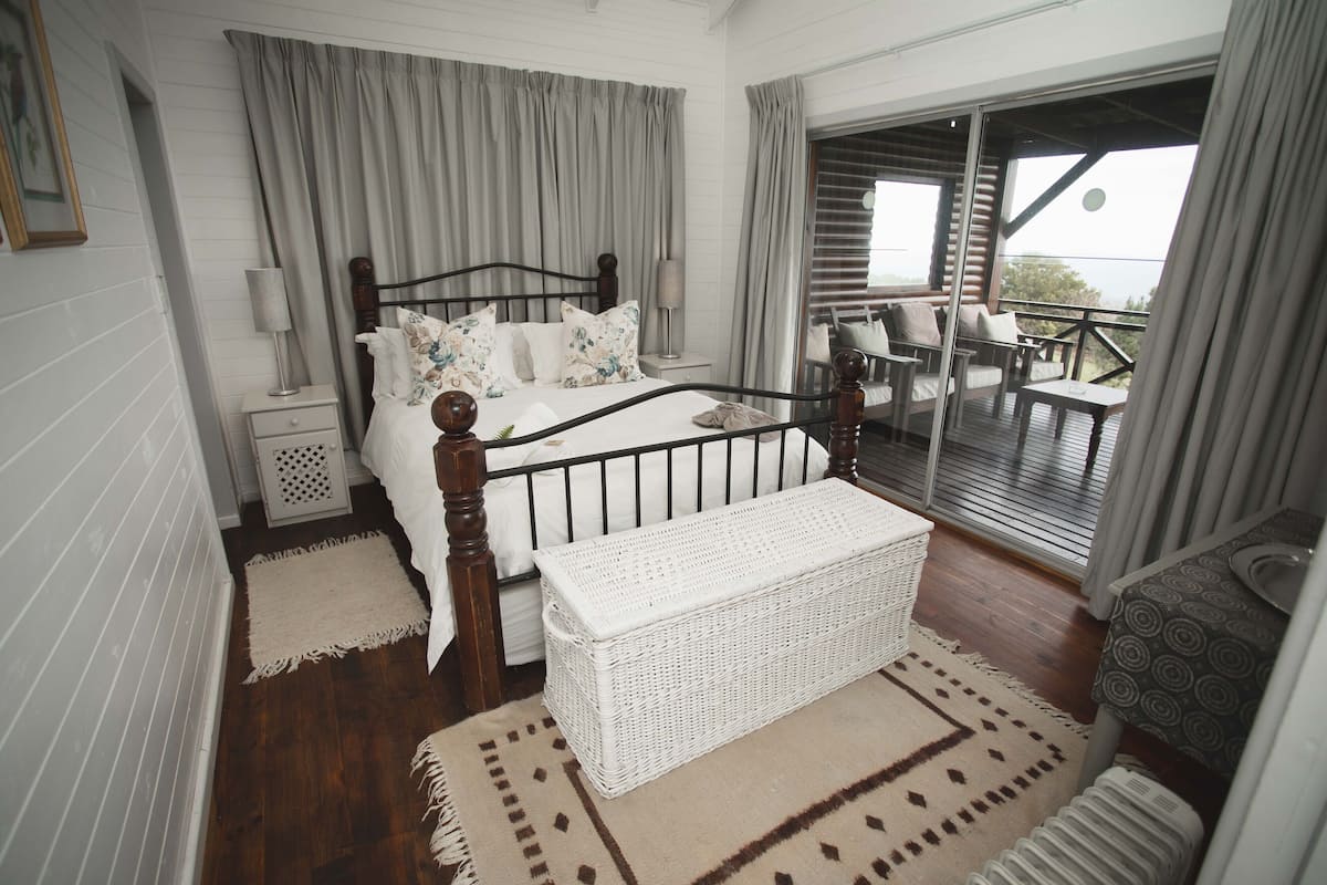 A bedroom with a wooden bed, a white bench, a balcony with outdoor furniture, and a view of the outdoors.