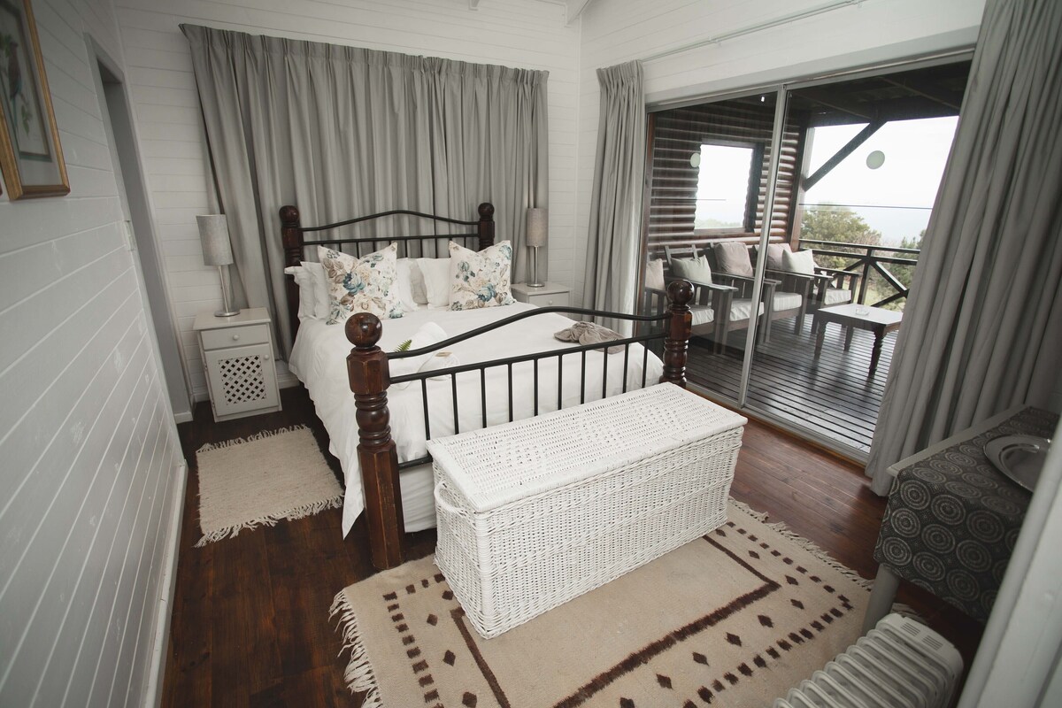 Un dormitorio con cama, mesitas de noche, un banco, un balcón con sillas y vistas.