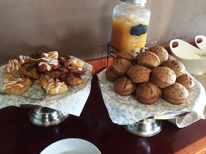 A breakfast buffet with pastries and muffins on marble trays, a glass jar of orange juice, and a white bowl.