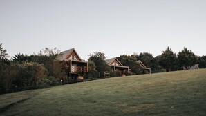 Un paysage rural avec deux maisons en bois sur une colline herbeuse, entourées d’une forêt dense.