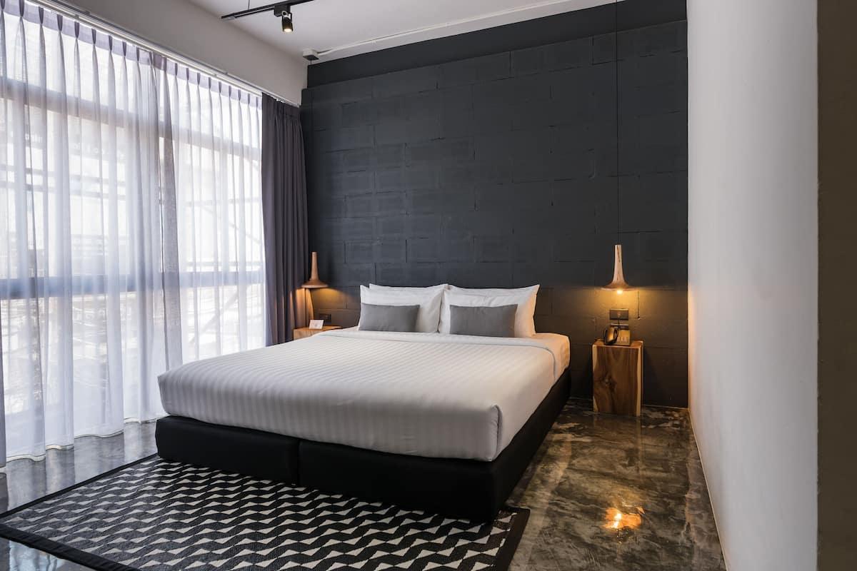A modern hotel room with a large bed, blackout curtains, a dark brick accent wall, bedside lamps, and a patterned rug.