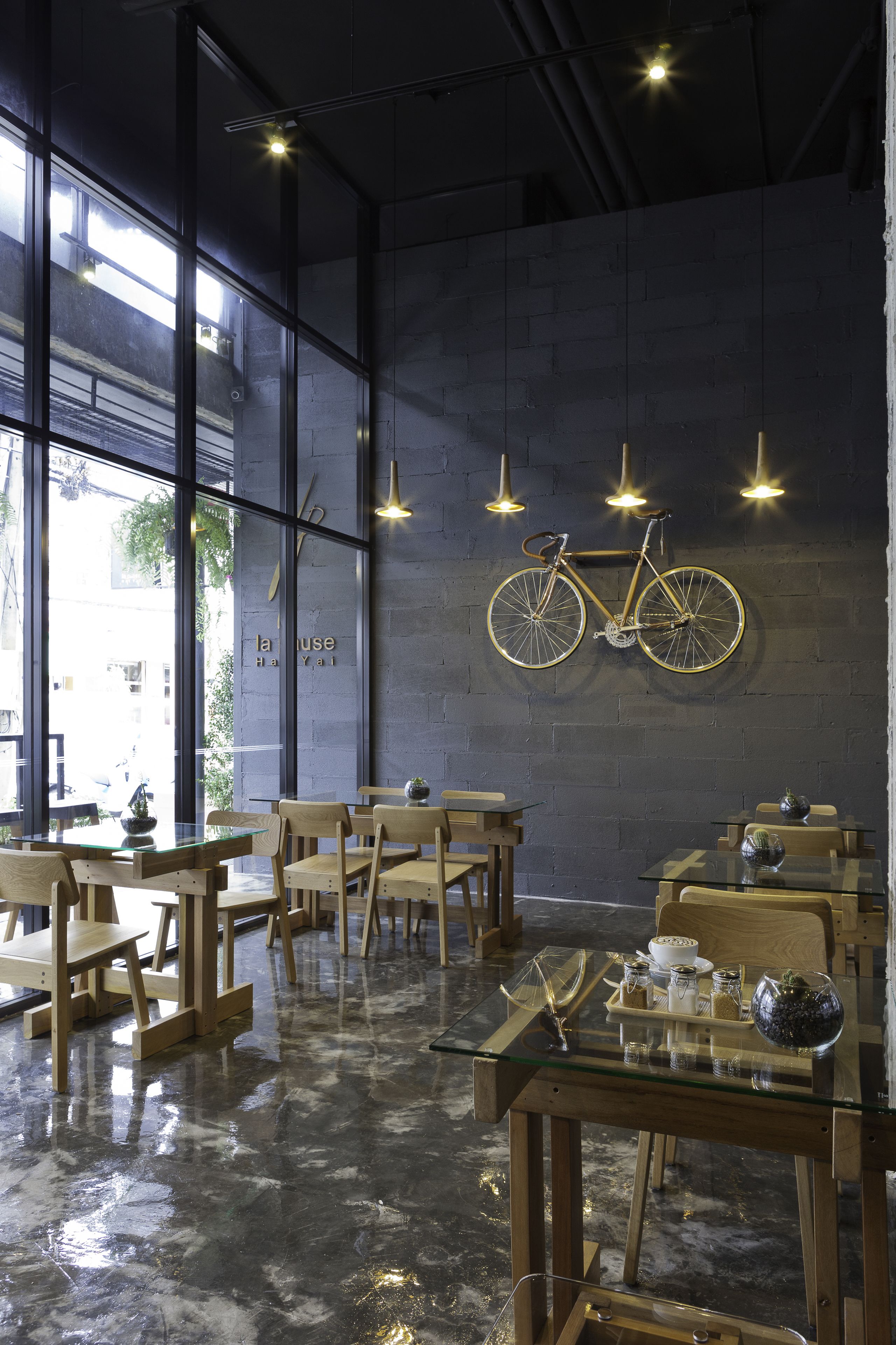 Et moderne caféinteriør med træborde og stole, en cykel ophængt på væggen, og store vinduer der lukker naturligt lys ind.