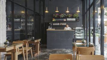 Interior d'una cafeteria amb taules i cadires de fusta, una barra amb una persona darrere i prestatges amb diversos objectes.