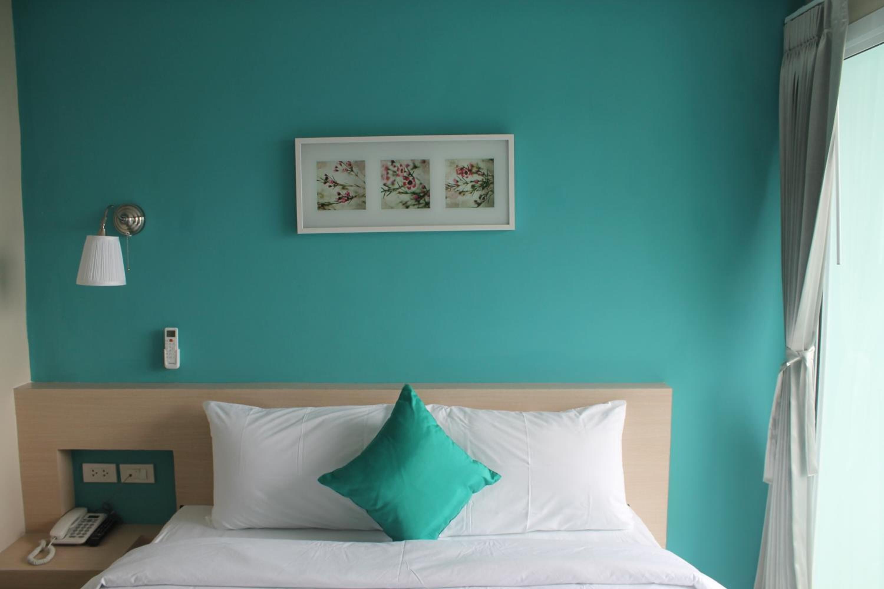 A hotel room with a teal wall, a bed with white bedding and a teal pillow, a bedside table with a phone, and a framed floral artwork above the bed.