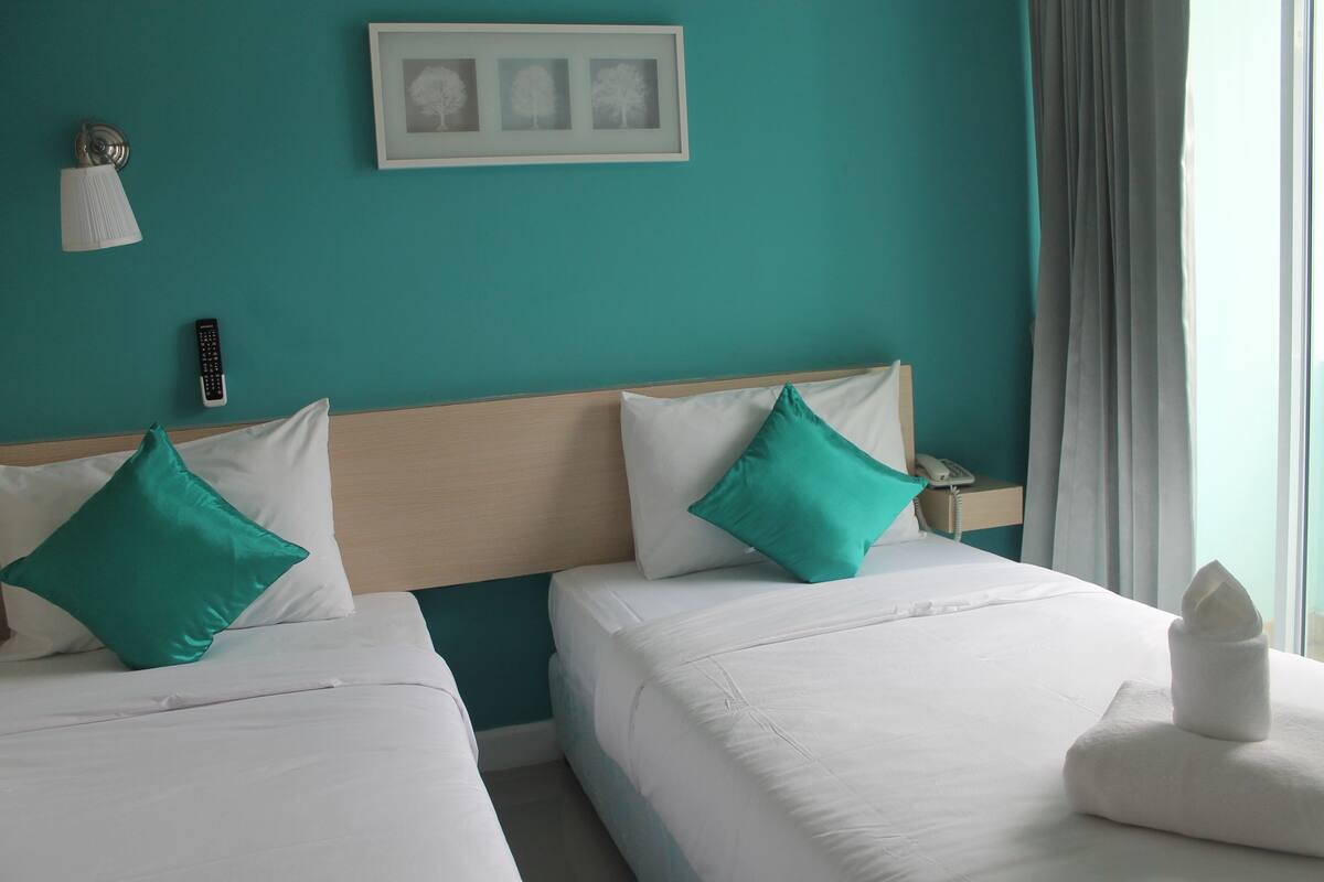 Habitación de hotel con dos camas, paredes verde azulado, un cuadro enmarcado, una lámpara y un teléfono en la mesita de noche.