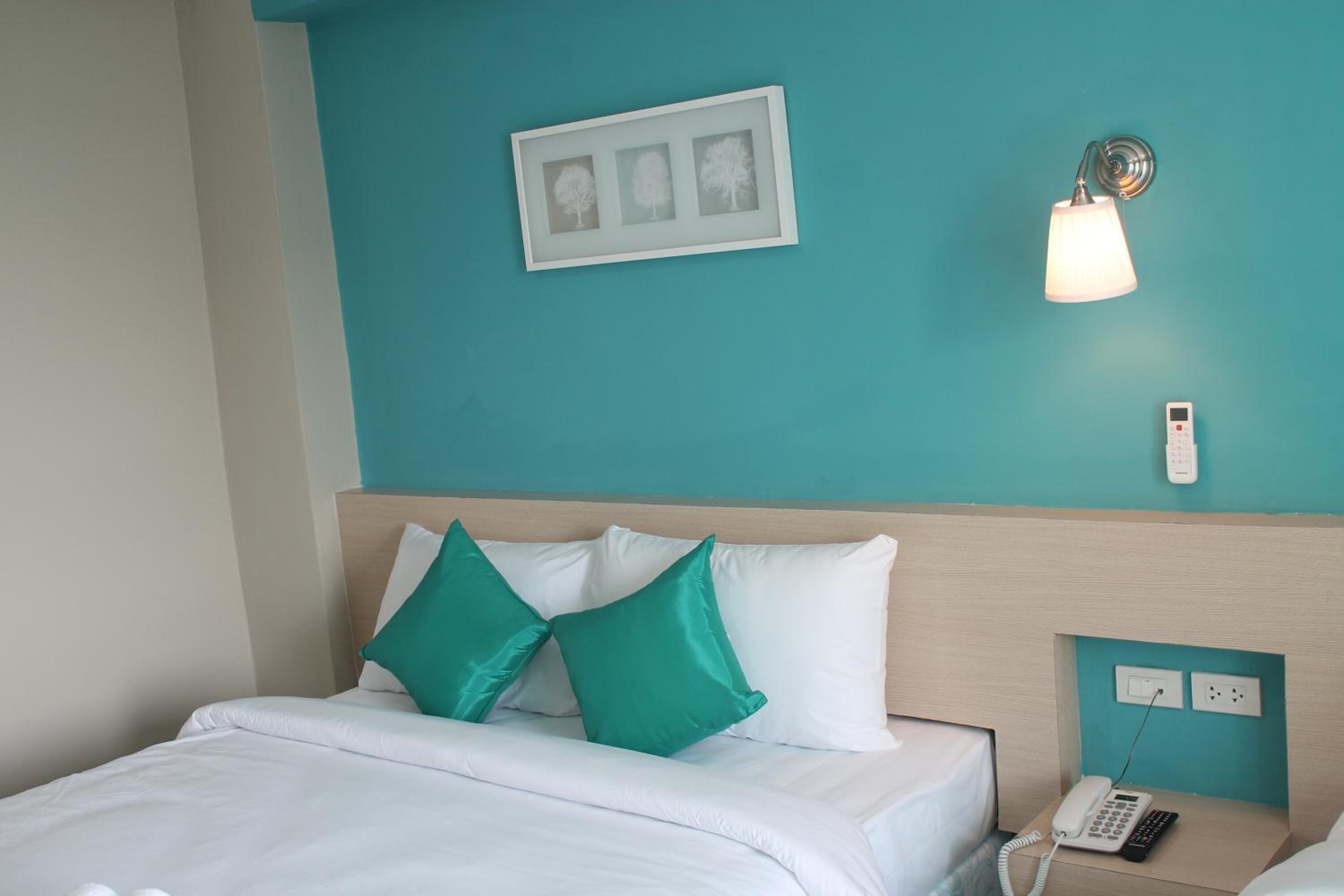 Habitación de hotel con pared verde azulada, cama con ropa blanca y dos almohadas verdes, mesita de noche con teléfono y lámpara, y un cuadro enmarcado sobre la cama.