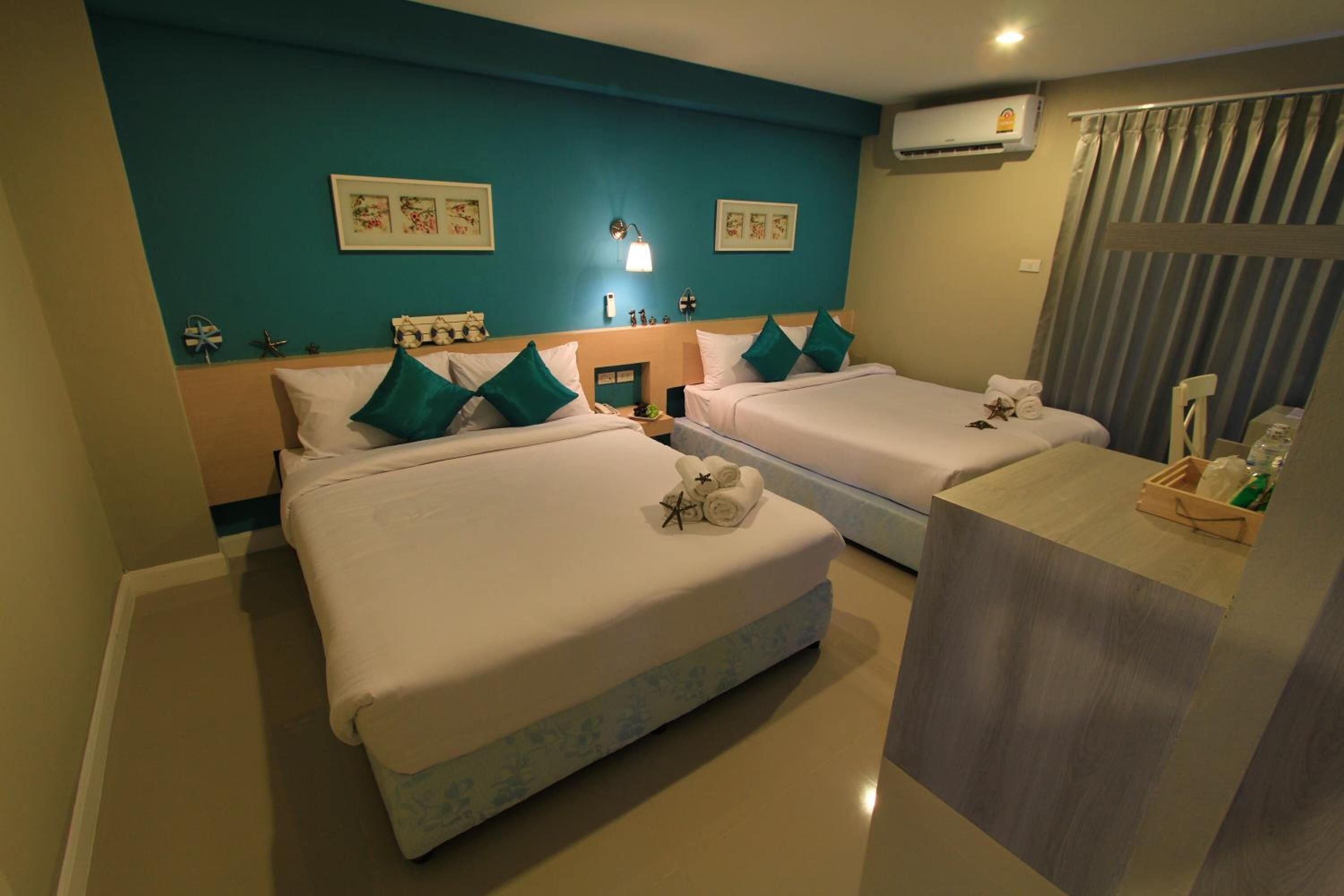 A hotel room with two beds, teal walls, a wooden headboard, a bedside table with a lamp, and a window with curtains.