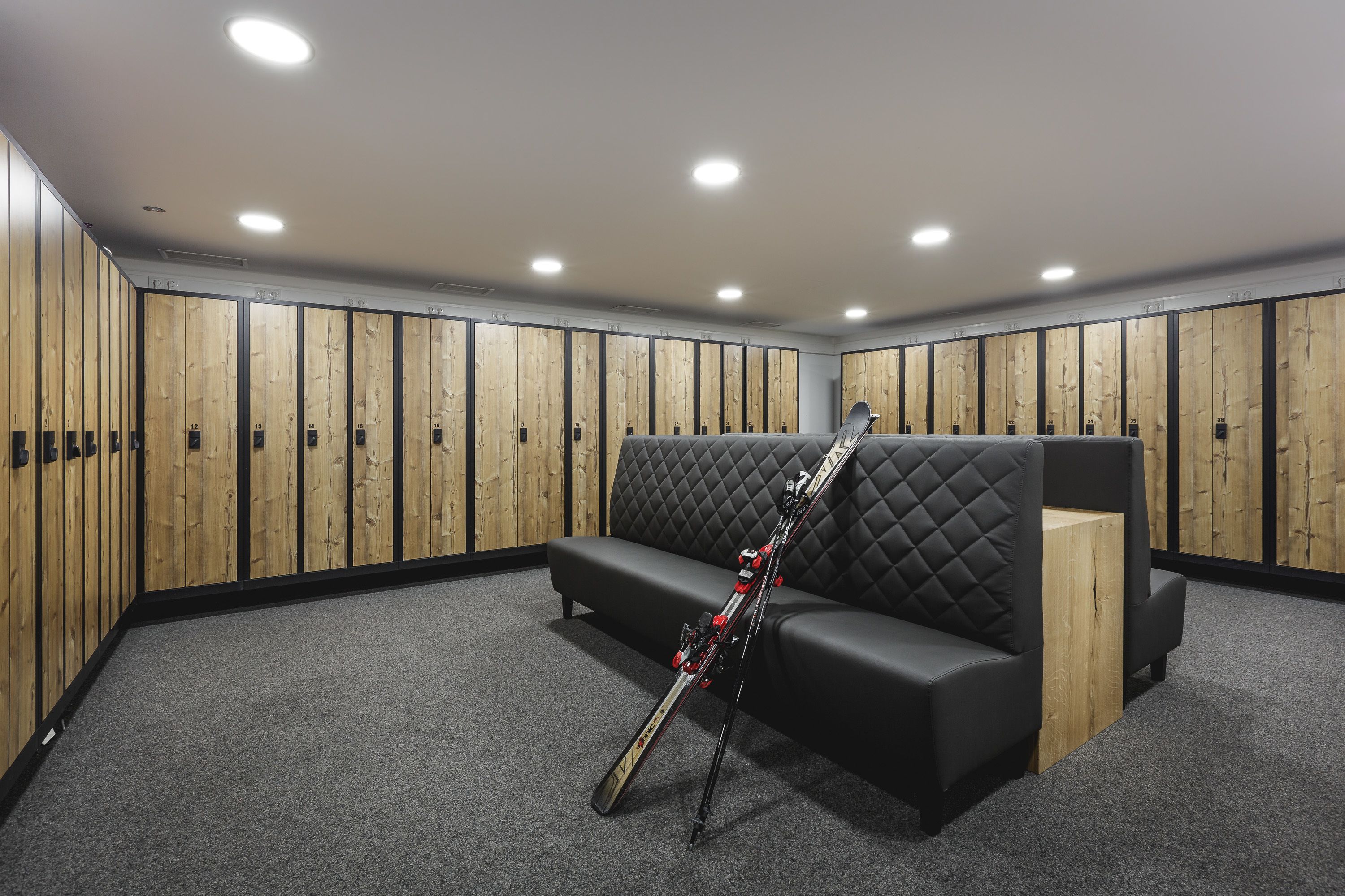 A locker room with wooden lockers, a black cushioned bench, and skis leaning against the wall.