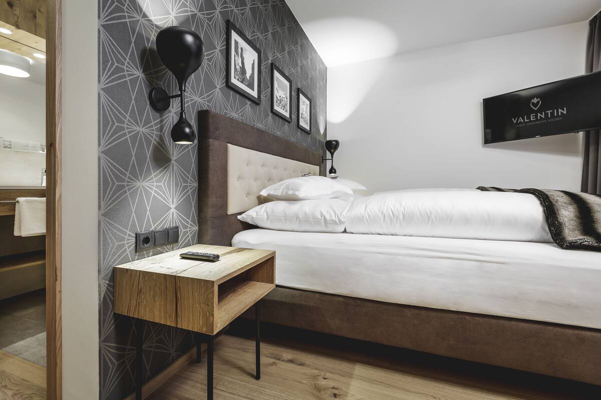 Un dormitorio moderno con una cama grande, una mesita de noche, un televisor montado en la pared y un papel tapiz con estampado geométrico.