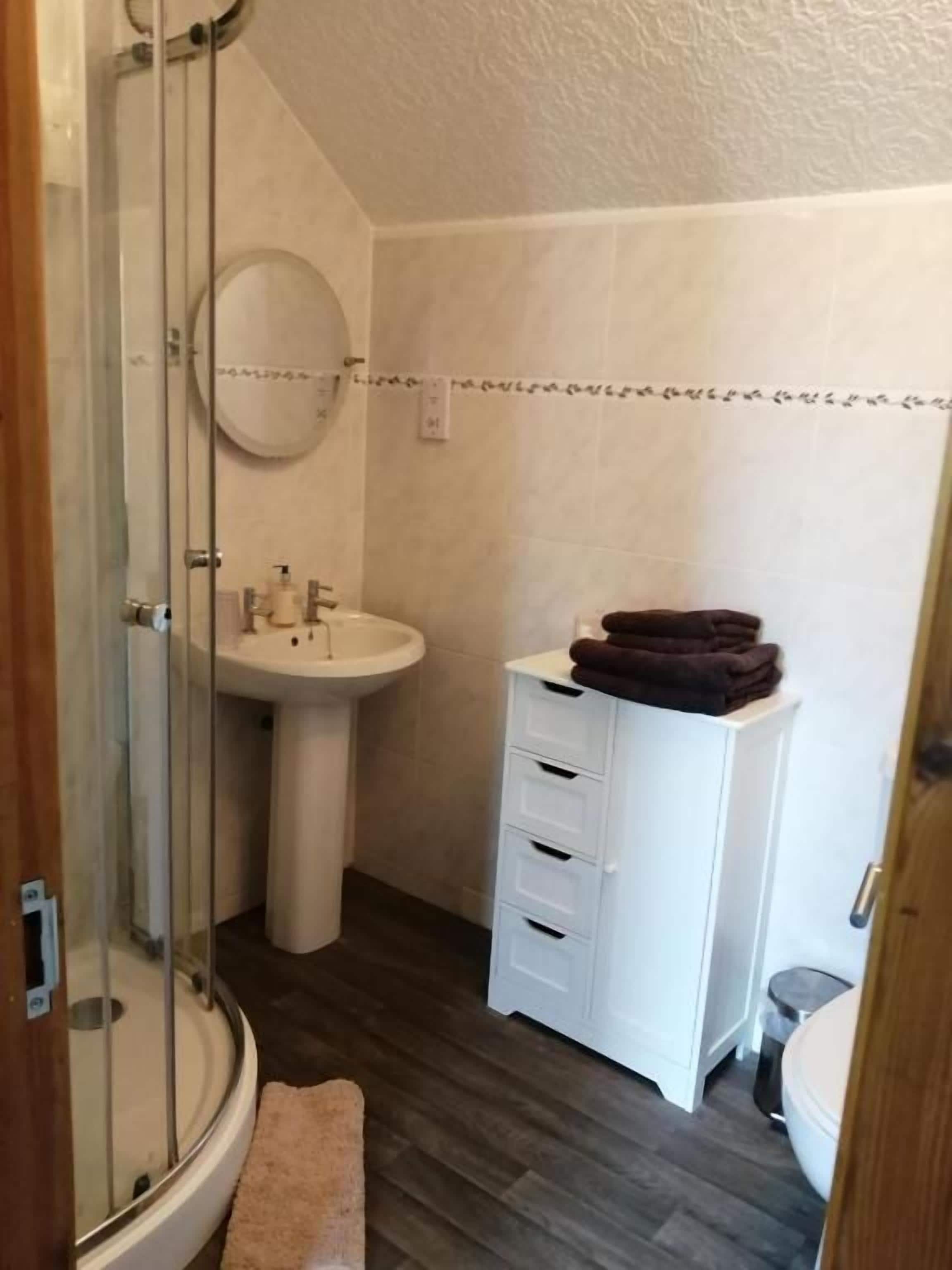A bathroom with a glass shower enclosure, a pedestal sink, a white vanity with drawers, and a toilet.