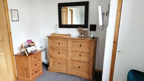 A room with a wooden dresser, a mirror, a blue chair, and a small shelf with items. - Don Muir Guest House (Oban)