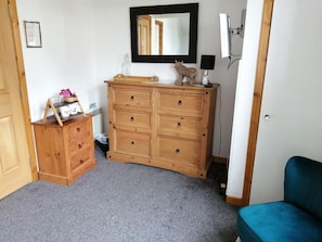A room with a wooden dresser, a mirror, a blue chair, and a small shelf with items. - Don Muir Guest House (Oban)