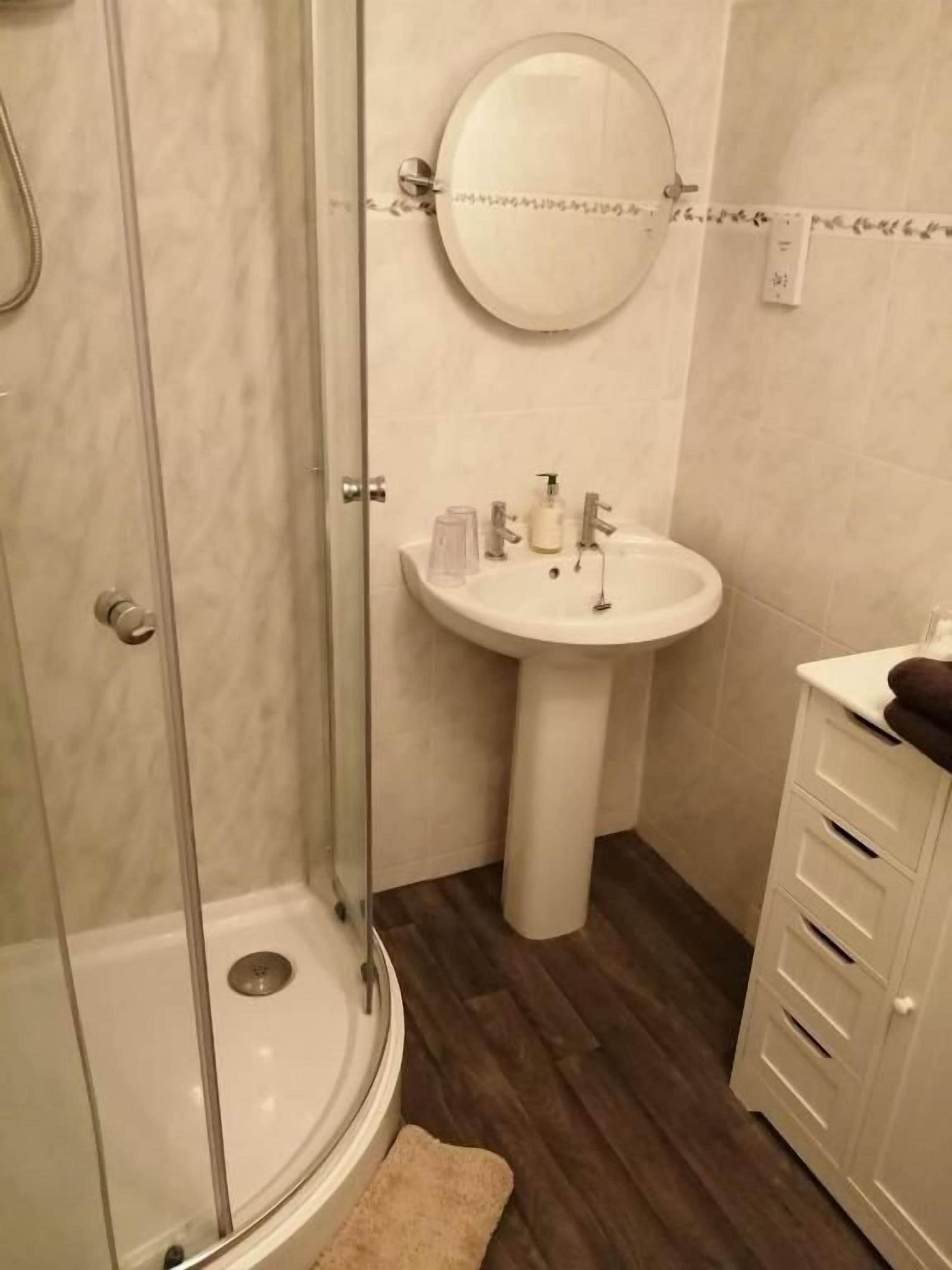 Un baño con ducha envuelta en vidrio, lavamanos sobre pedestal y un armario.