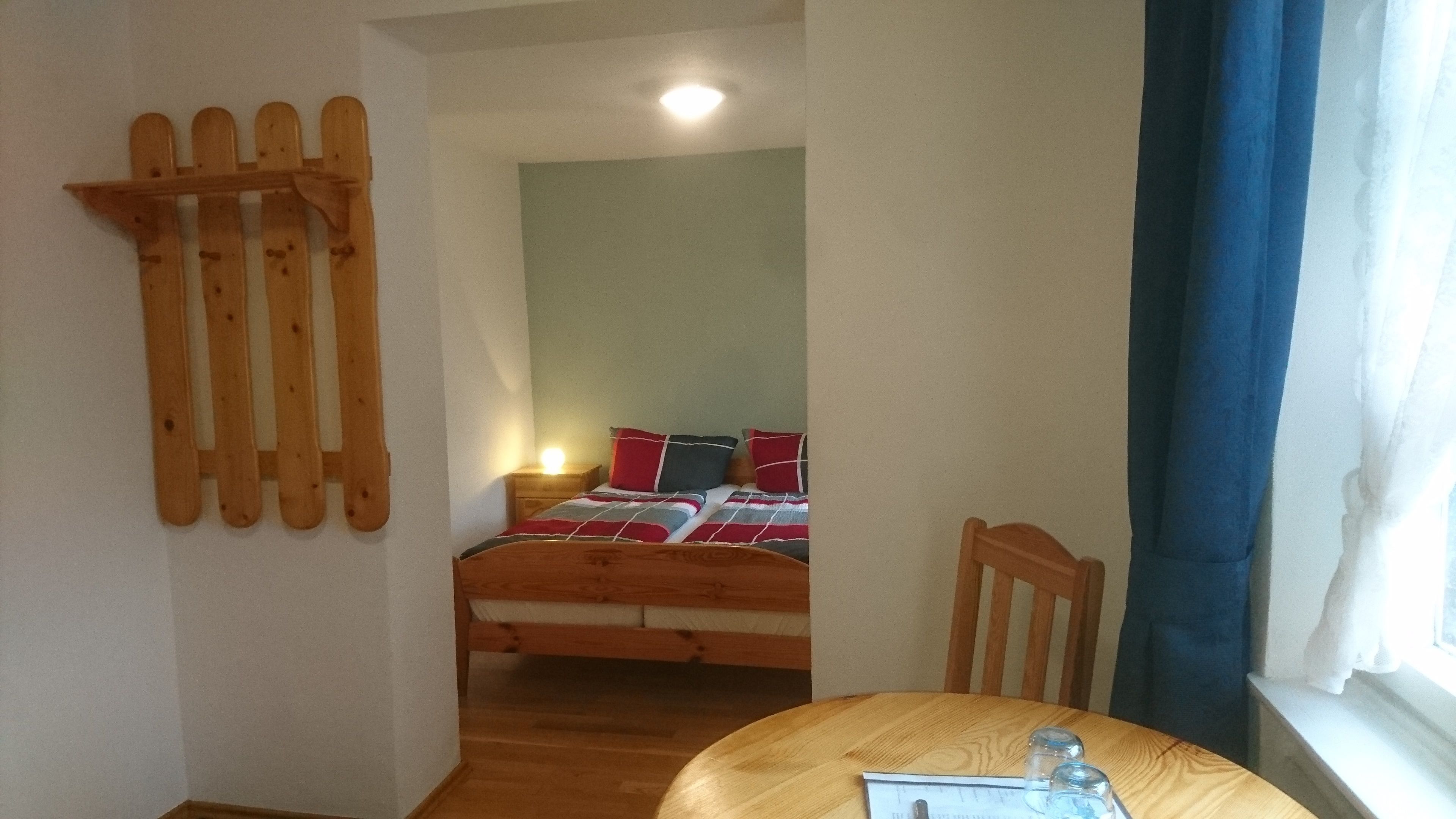 A small, cozy room with a wooden bed, a nightstand, a table, and a window with curtains.