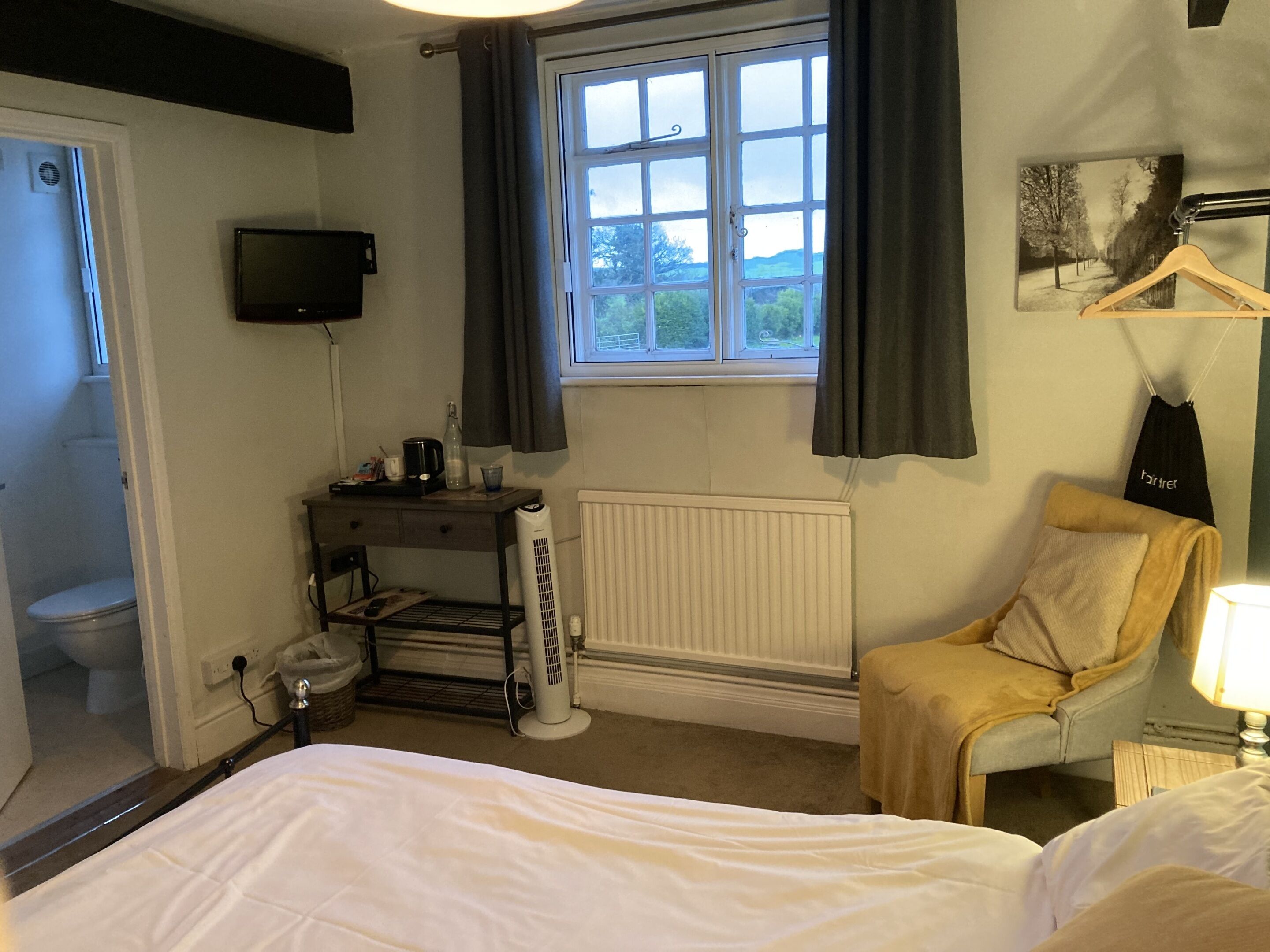 A hotel room with a bed, a chair, a desk with a computer, a radiator, a window with curtains, a television, and a picture on the wall.