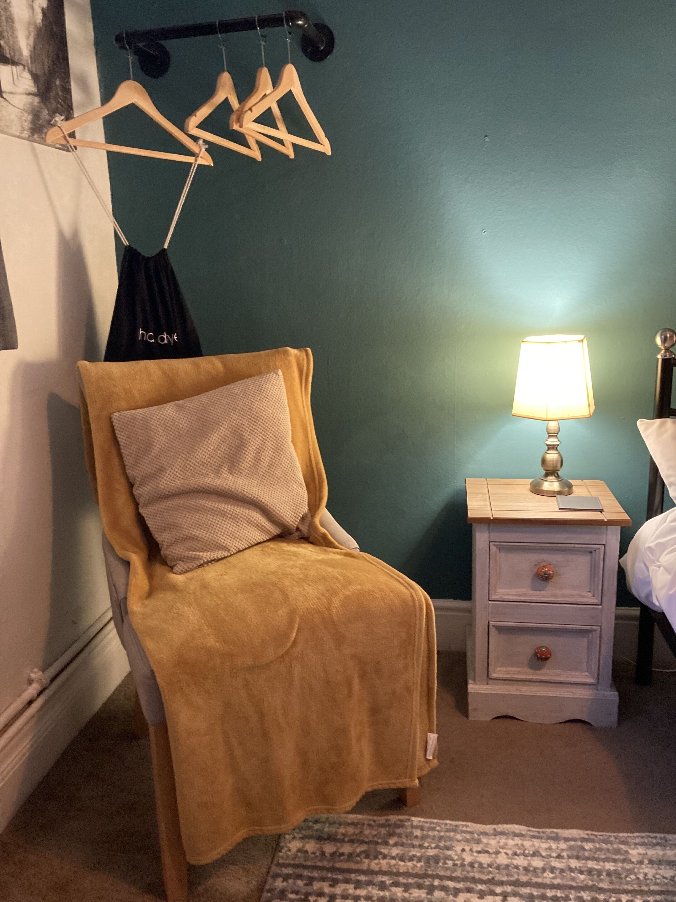 A bedroom with a single bed, a bedside table with a lamp, a chair with a blanket, and a wall-mounted coat rack.