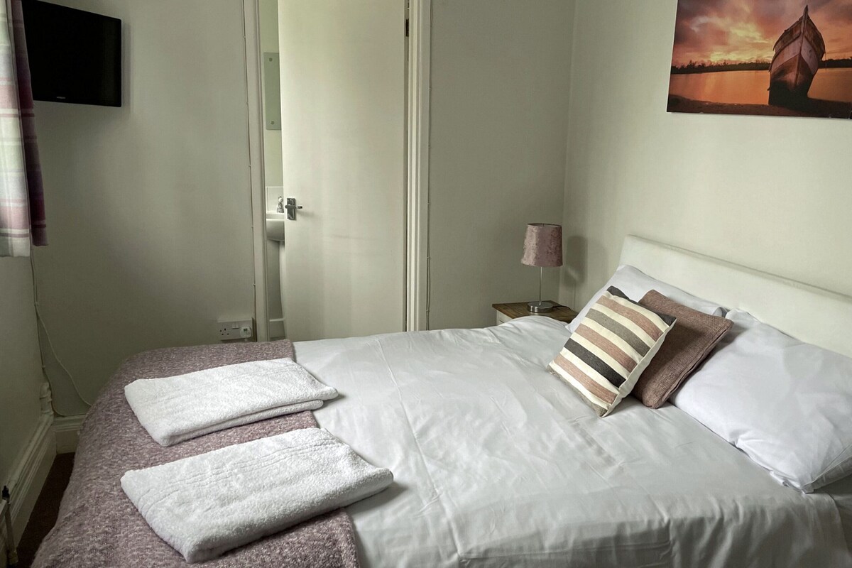 A neatly made bed with white linens and striped pillows, a bedside lamp, and a painting of a boat on water.