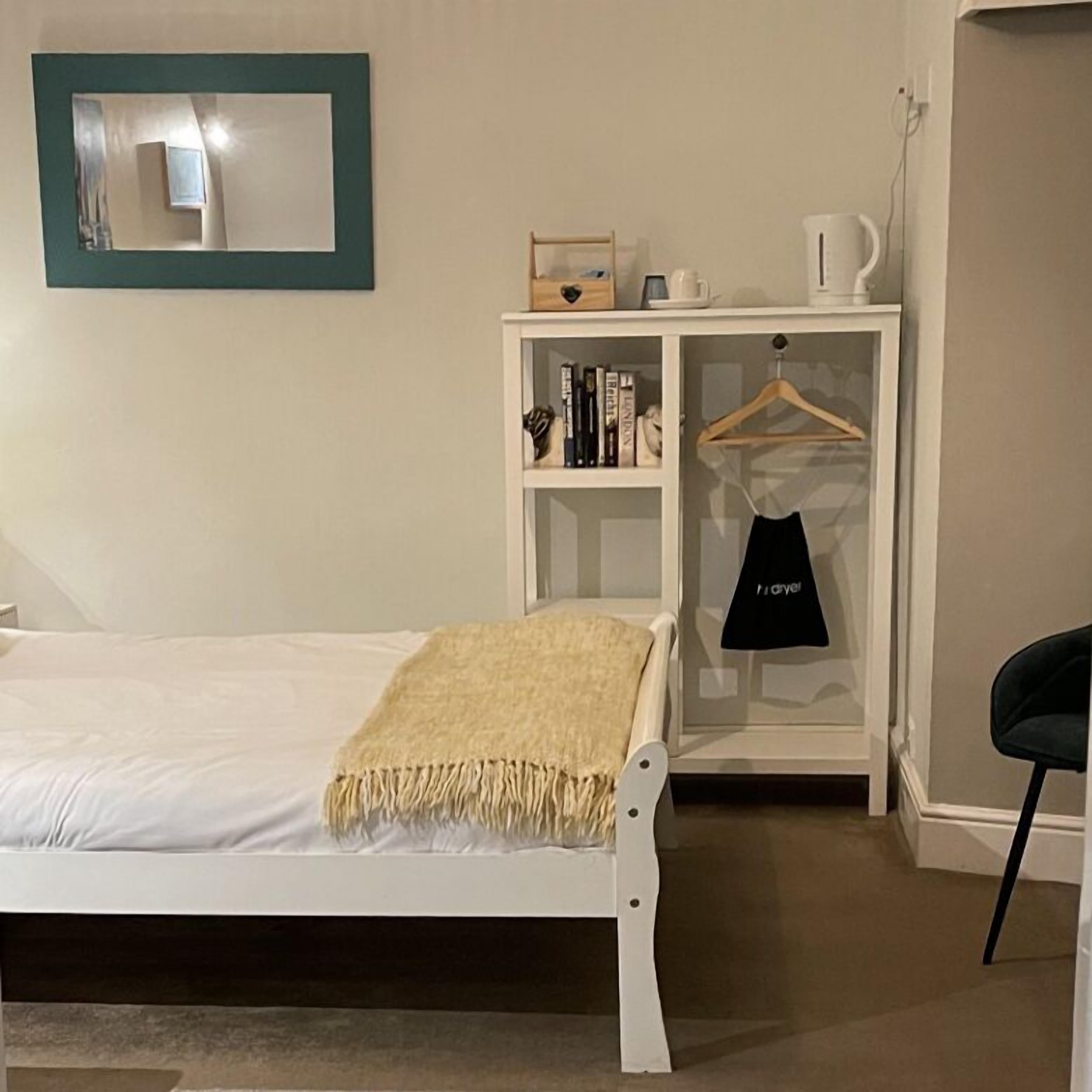 A neatly arranged bedroom with a bed, a bedside table with a lamp, a hanging garment rack, and a small cabinet with books.
