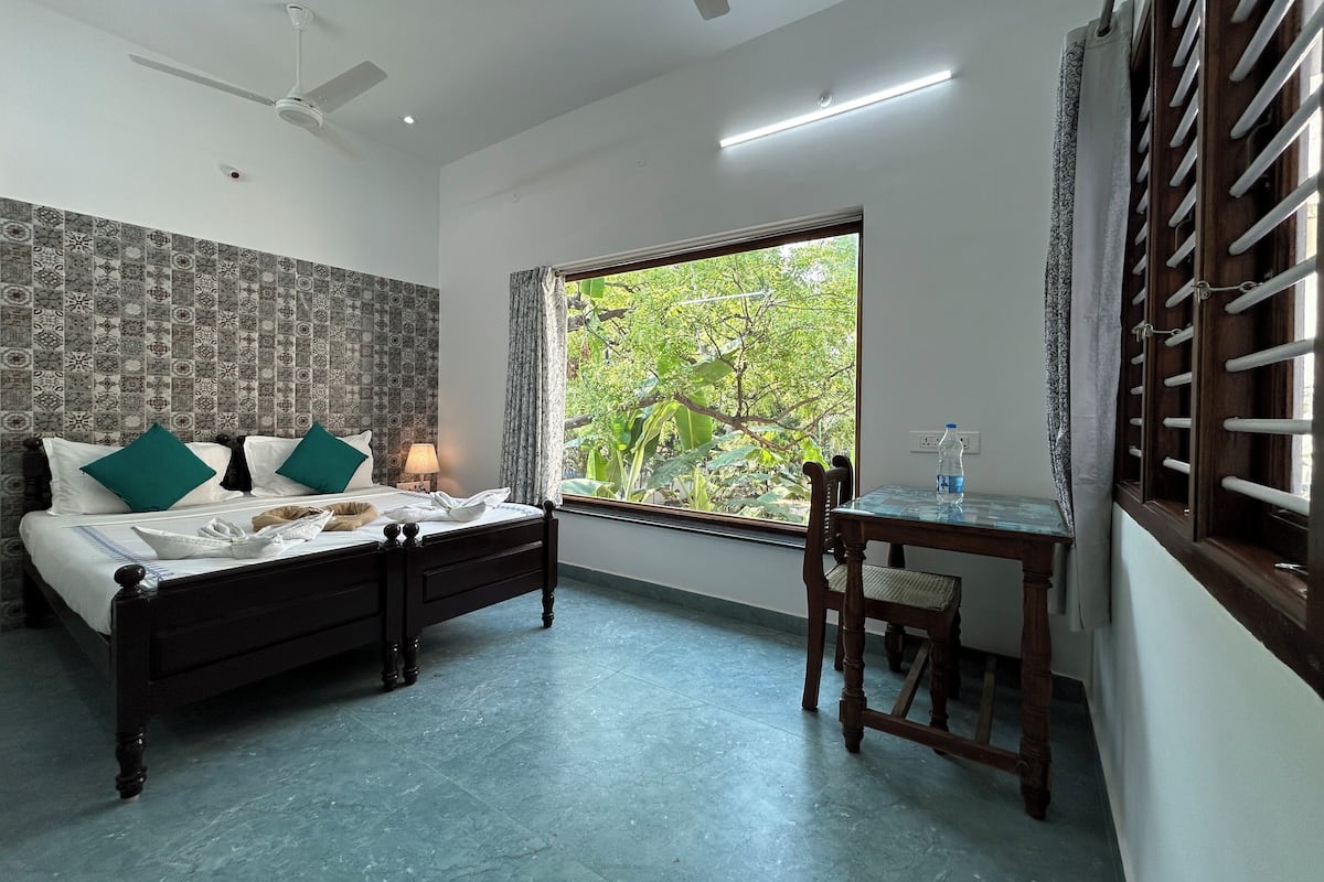 A bedroom with a bed, a desk, a chair, a ceiling fan, and a window with a view of greenery.