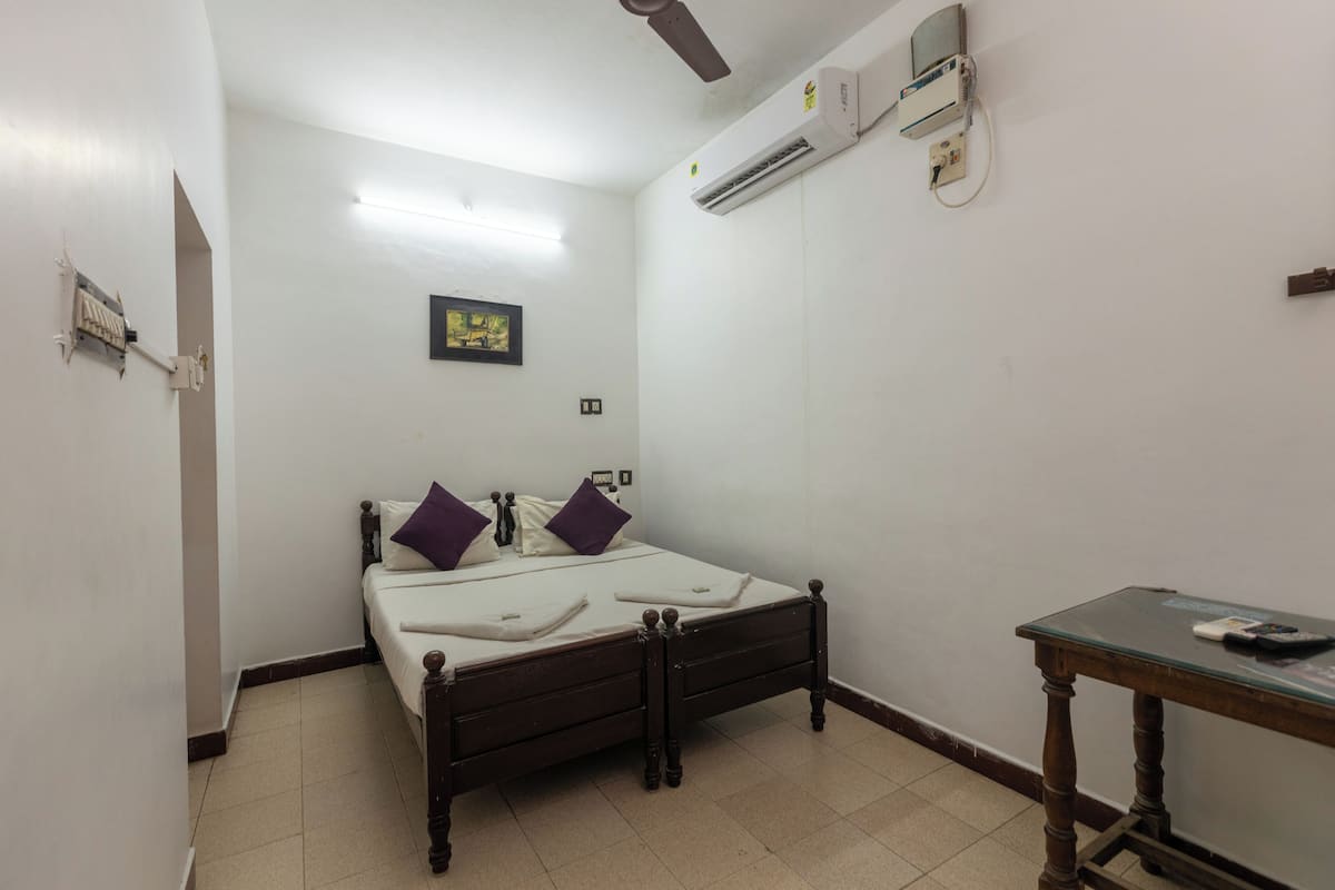 A small, narrow room with a single bed, a wooden table, and an air conditioning unit mounted on the wall.
