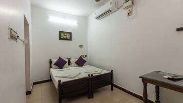A small, narrow room with a single bed, a wooden table, and an air conditioning unit mounted on the wall.