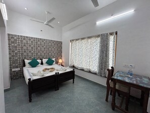 A hotel room with a bed, a desk, a chair, a ceiling fan, a window with curtains, and a wall with patterned tiles. - Hotel Coramandal Heritage (Puducherry)