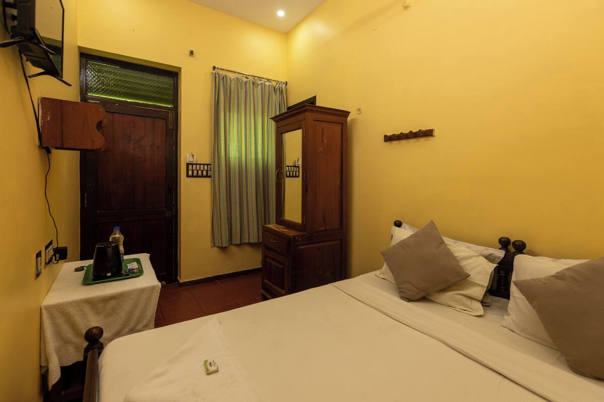 A hotel room with a bed, a wooden cabinet, a small table with a kettle, and a curtain.
