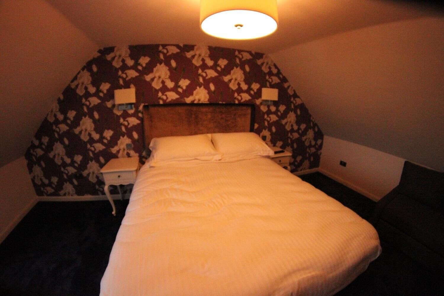 Una habitación de hotel con una cama grande, un cabecero estampado con motivos florales, dos mesitas de noche y una lámpara de pared.