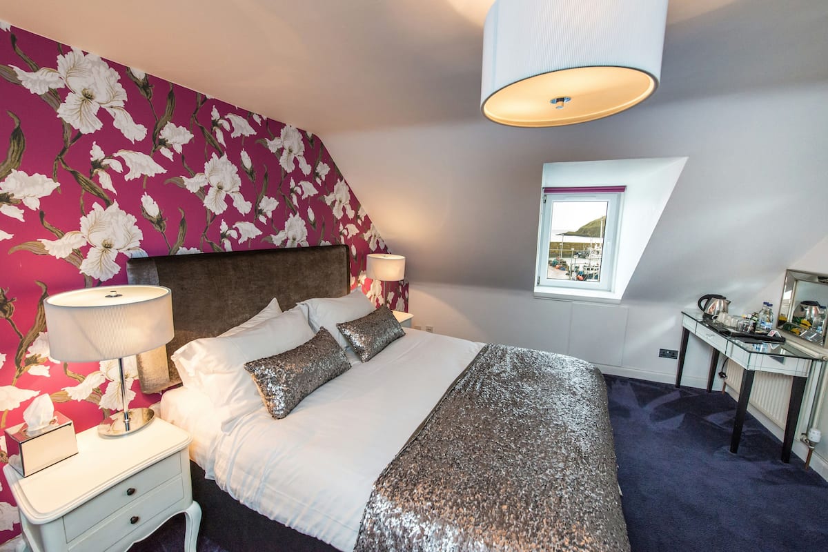 A bedroom with a bed, nightstands, a lamp, a window with a view, and a desk with items on it.