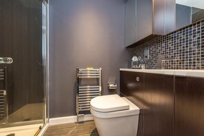 Un baño con cabina de ducha de vidrio, inodoro blanco, toallero y tocador de madera oscura con salpicadero de azulejos.
