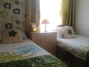 A hotel room with two beds, a bedside table with a lamp, and a window with curtains.