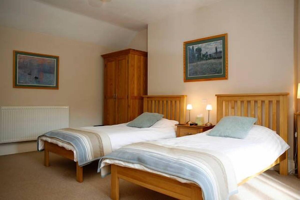 A bedroom with two single beds, wooden headboards, bedside tables with lamps, and two framed pictures on the walls.
