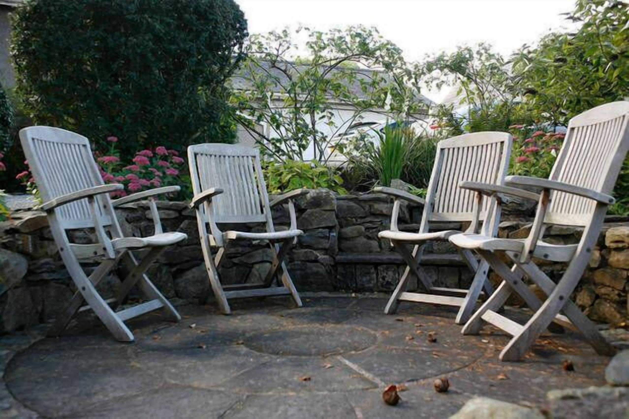 Trois chaises pliantes blanches disposées en demi-cercle sur une terrasse en pierre, avec un mur de pierre et de la verdure en arrière-plan.