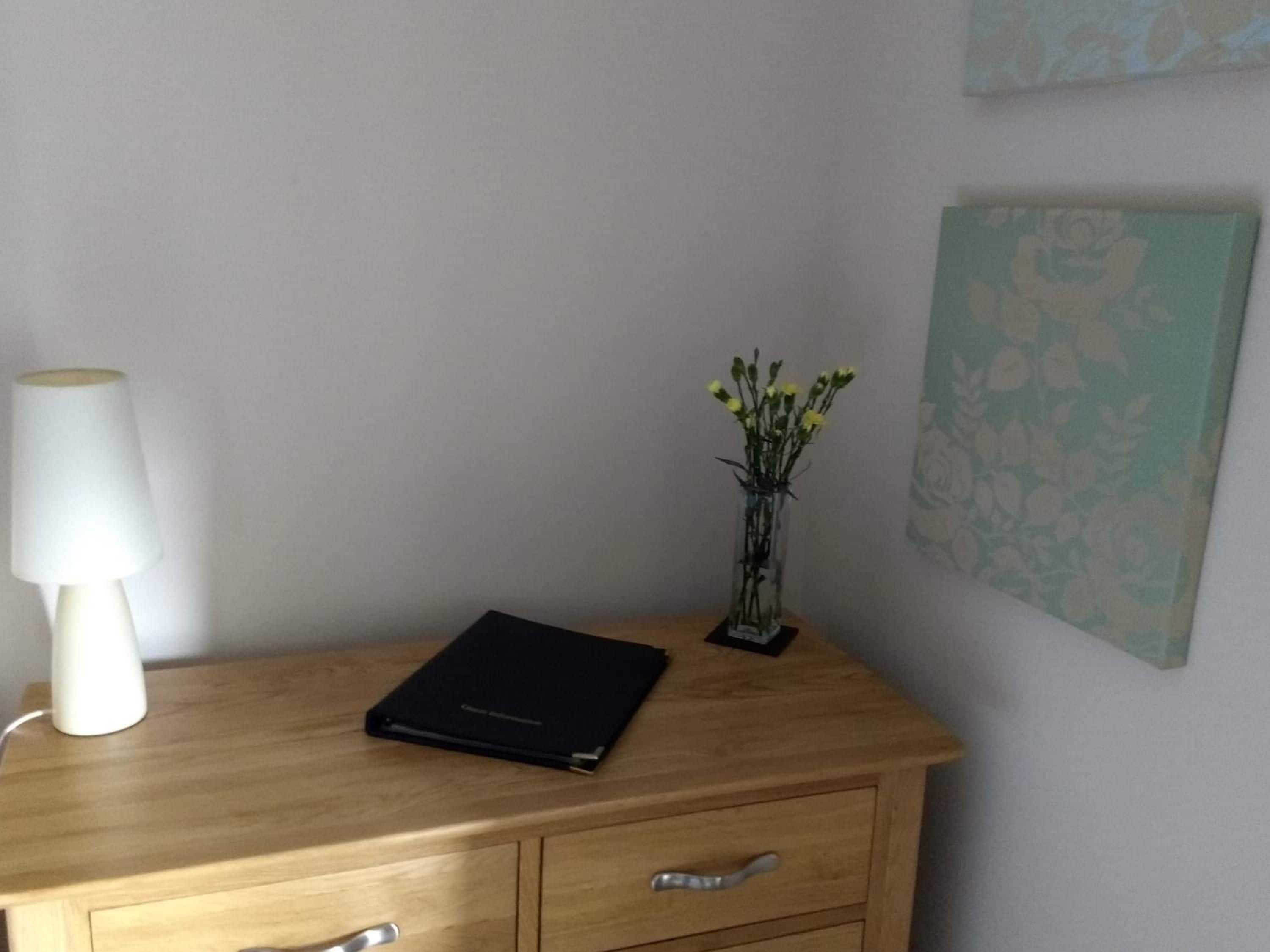 Uma mesa de madeira com abajur, um vaso com flores e um quadro na parede.