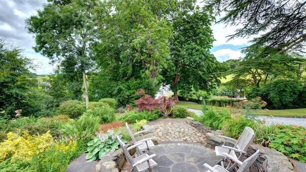 A stone patio with outdoor furniture, surrounded by lush greenery and a small pond.