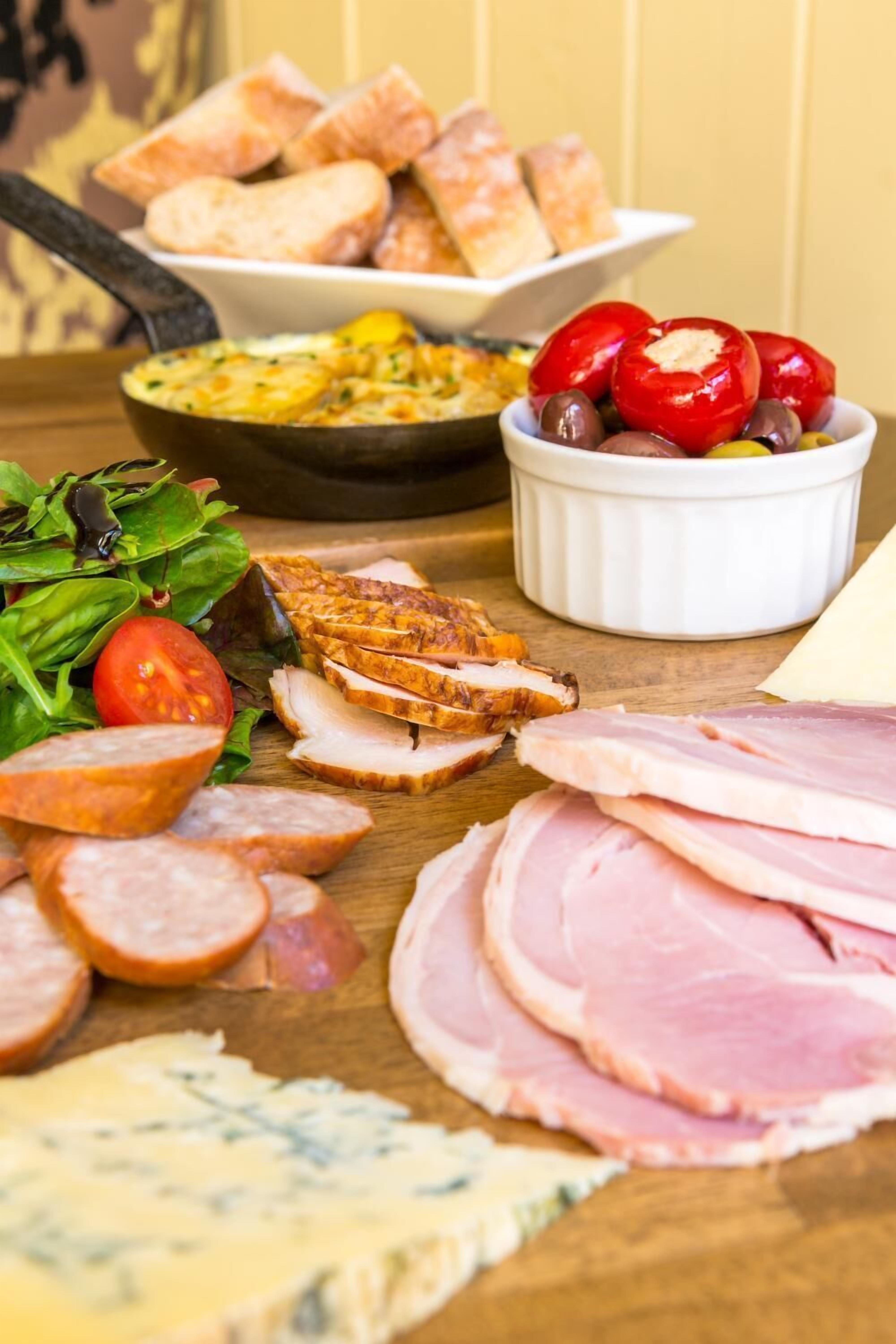 Diversos alimentos, incluyendo jamón en rodajas, salchichas, queso y un bol de aceitunas y tomates cherry.