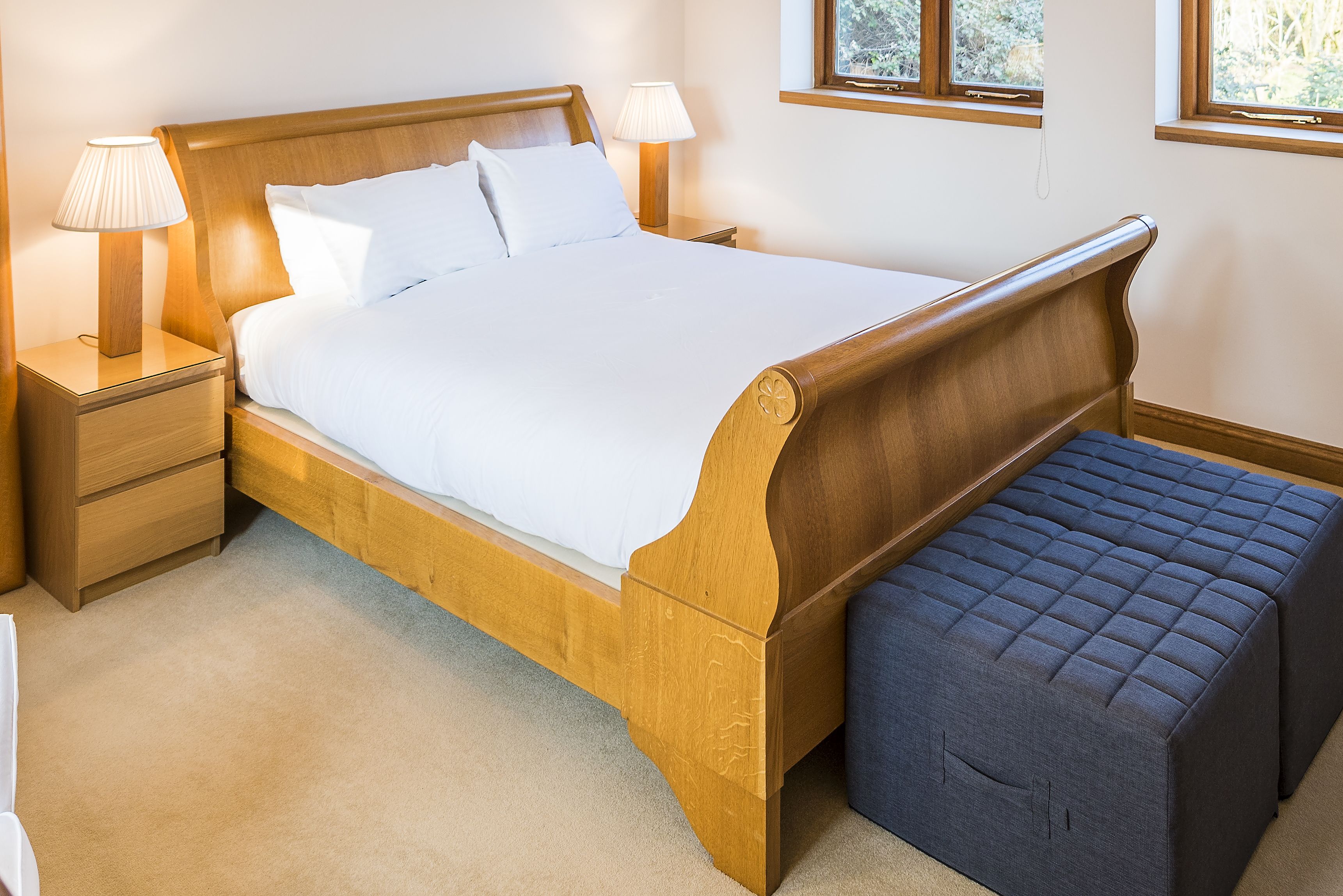 Una cama de madera con ropa de cama blanca, dos lámparas de noche y un banco.