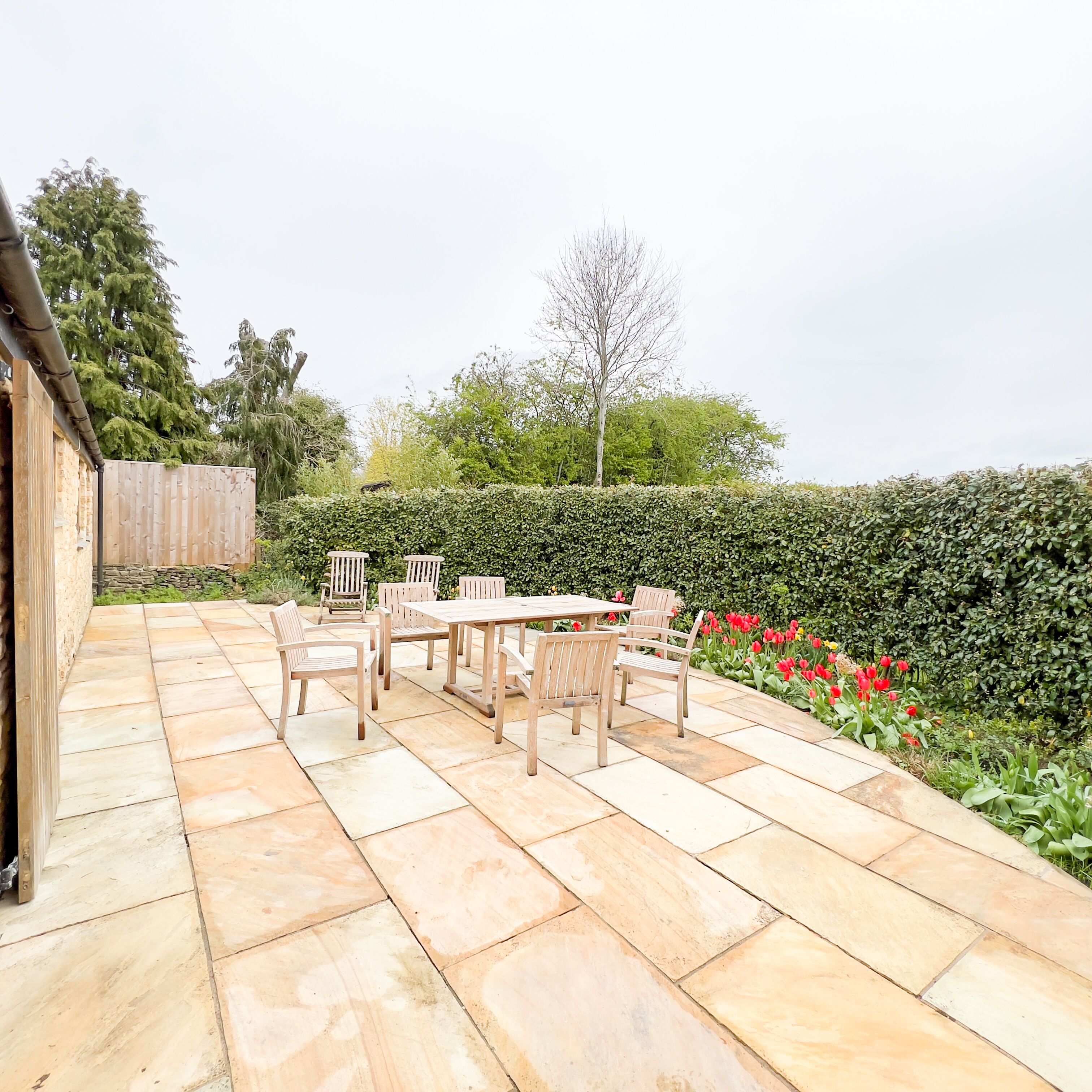 A patio with a wooden table and chairs, surrounded by a hedge and red flowers.