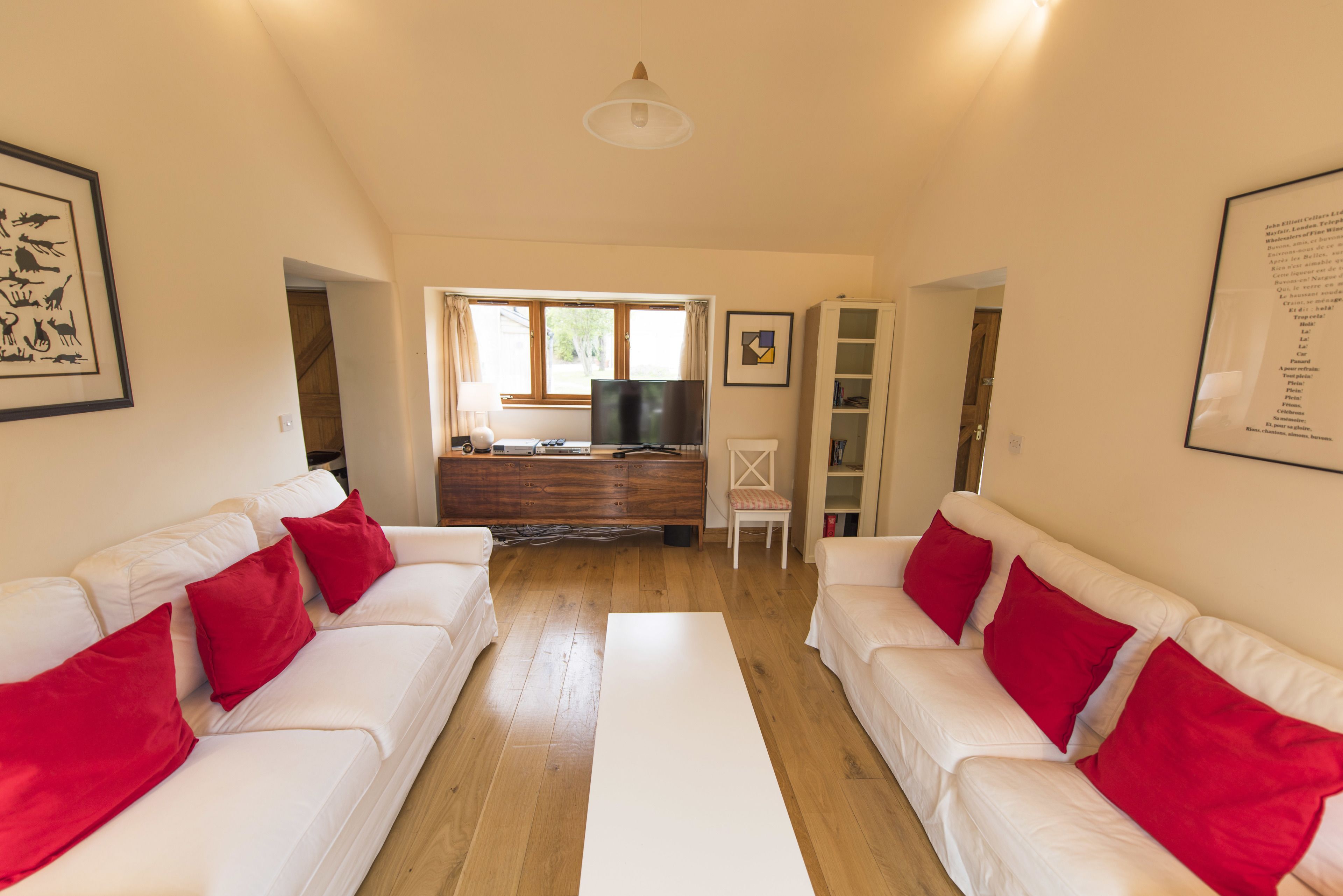 A living room with two white sofas, red cushions, a flat-screen TV, wooden flooring, and a large window.