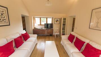 A living room with two white sofas, red cushions, a flat-screen TV, wooden flooring, and a large window.