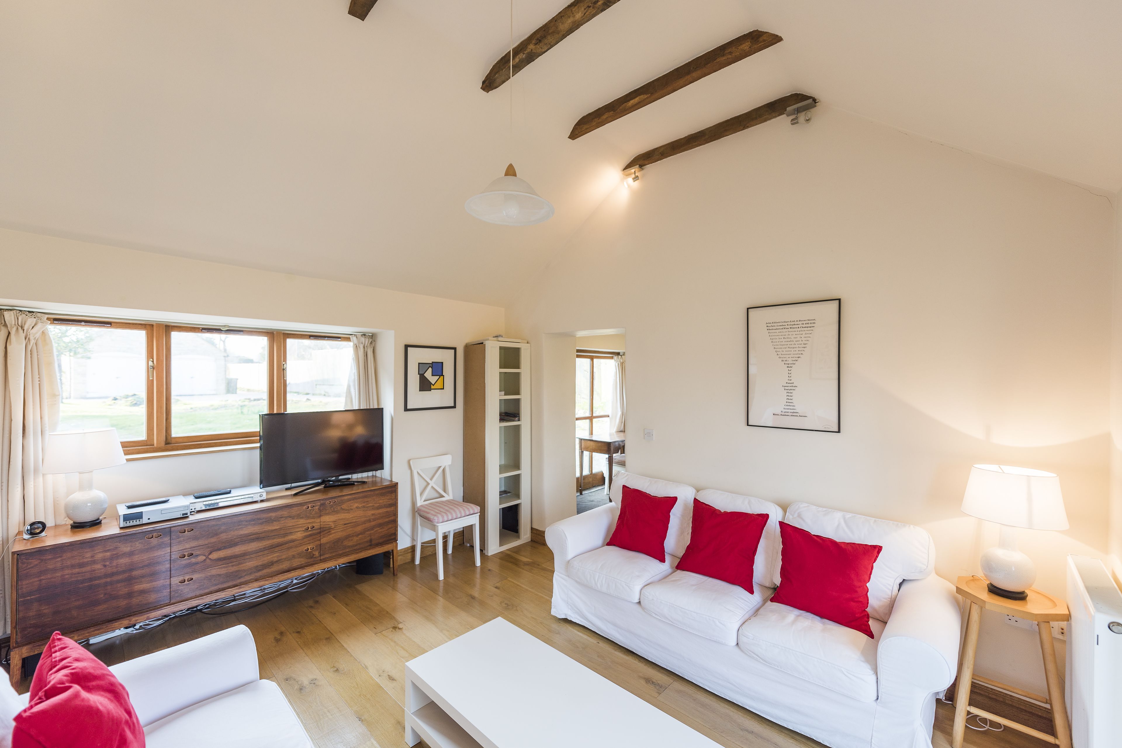 Een moderne woonkamer met een witte bank, rode kussens, een flatscreen televisie, houten balken in het plafond en een groot raam met gordijnen.
