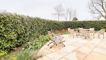 An outdoor patio with a wooden table and chairs, surrounded by a hedge and flowering plants.